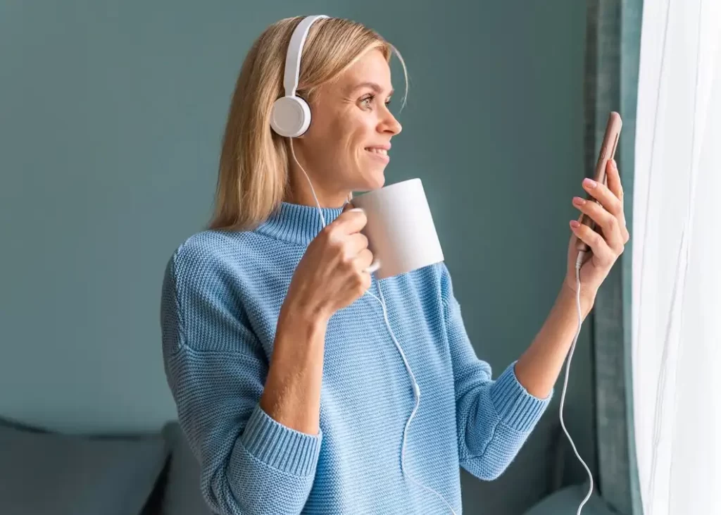 Mulher ouvindo música com fones de ouvido, segurando uma caneca enquanto verifica o celular, ideal para Daily Listen.